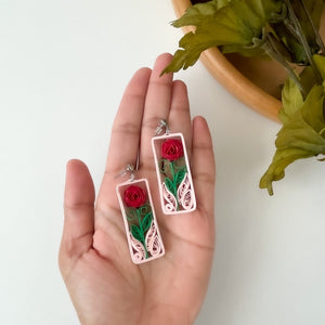 Handmade earrings with a paper quilled red rose and leaves, captured in a light pink frame.