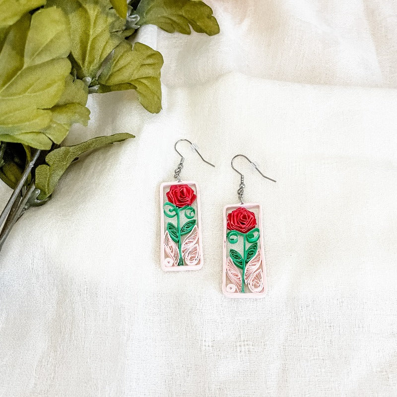 Handmade earrings with a paper quilled red rose and leaves, captured in a light pink frame.